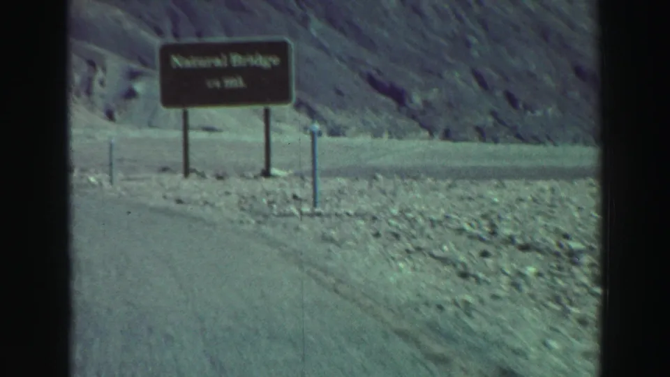 Closer 1969 8mm view of the Natural Bridge roadside sign near a curve through rocky Lake Tahoe-area terrain.