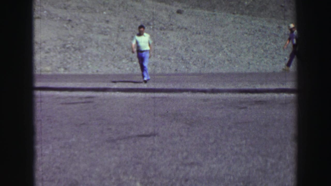 Two men walking near a paved path below a barren gray slope.