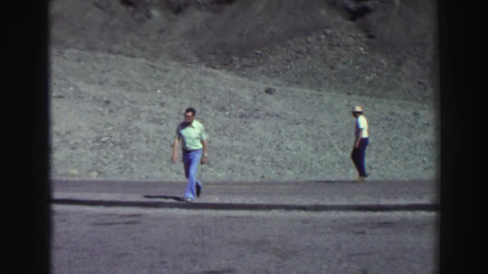 1969 Lake Tahoe frame with one man in a light green shirt walking in front of a gray slope and another man farther back.