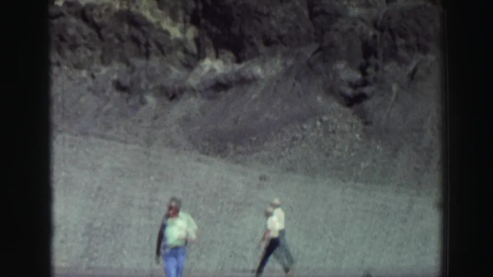 1969 Lake Tahoe home-movie frame with two adult men walking below a rocky gray mountain face, partly softened by motion blur.