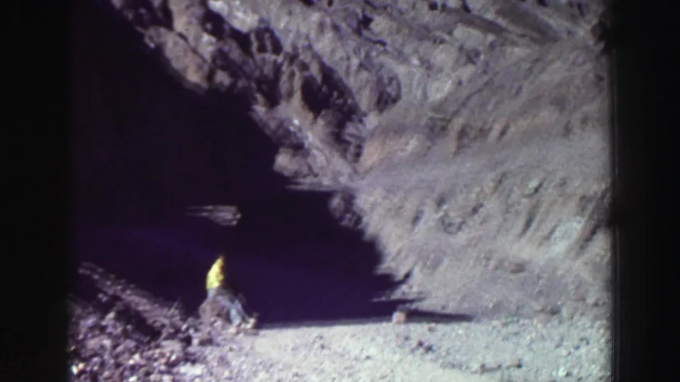 1969 Lake Tahoe frame with a distant person in yellow beside a dark cave entrance and sunlit rock walls.