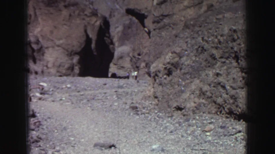 1969 Lake Tahoe home-movie frame showing a dark cave opening, rough rock face, pale ground, and a small distant visitor.