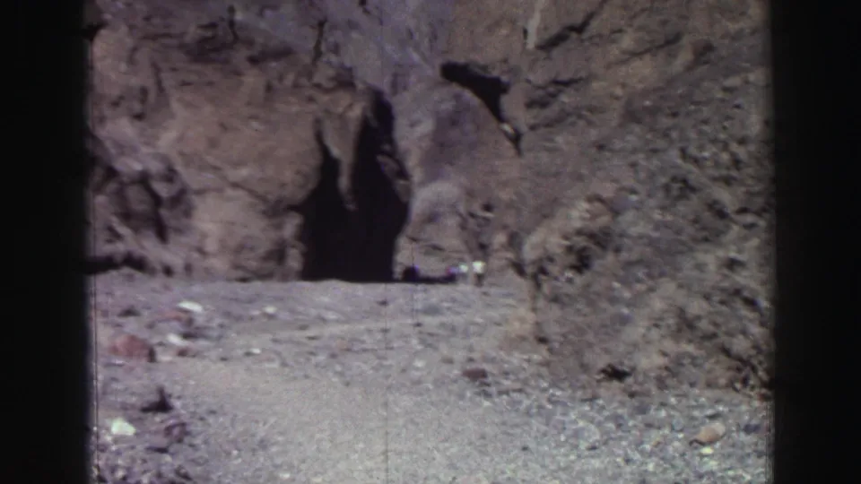 1969 Lake Tahoe frame showing a shaded cave entrance, rocky foreground, and a distant visitor partly visible near the opening.
