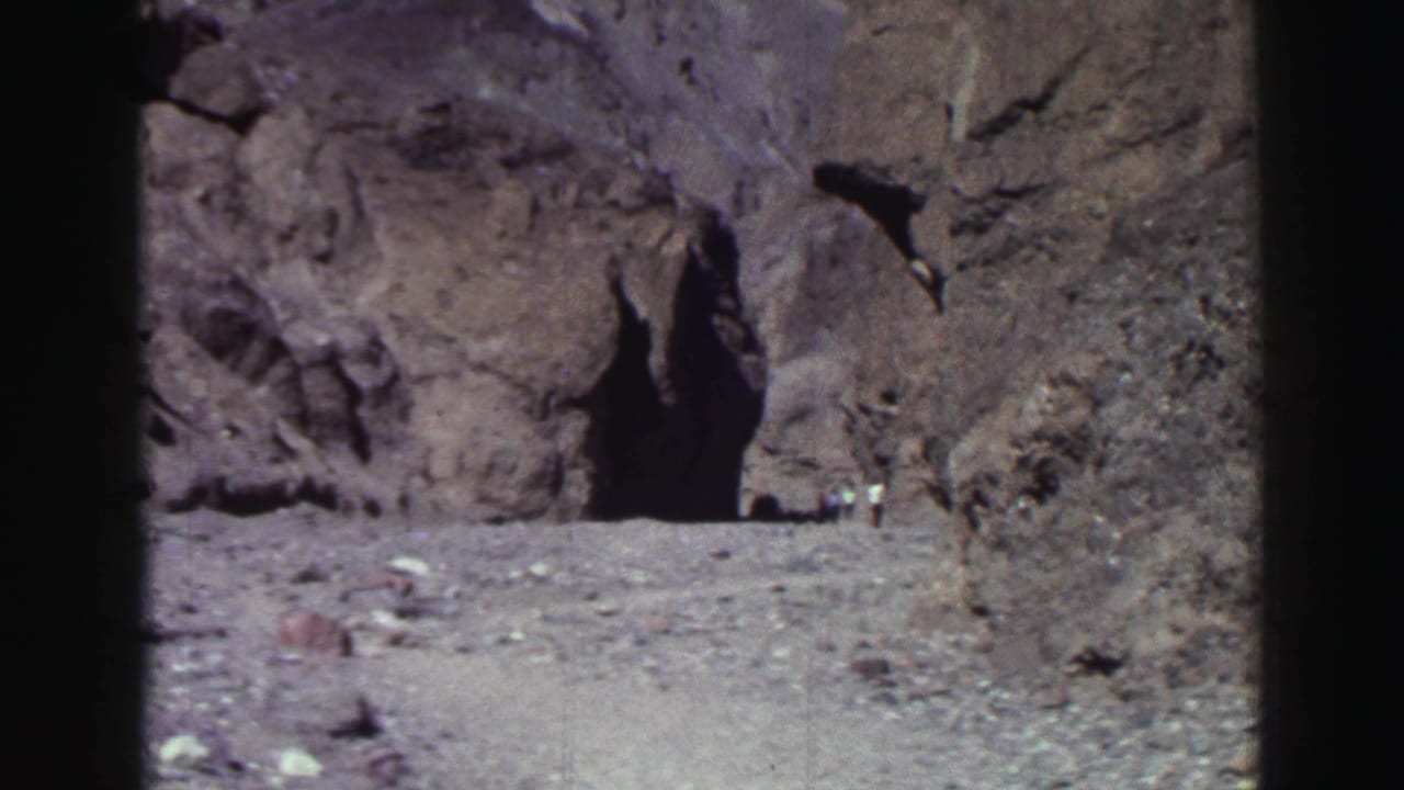 Rocky cliff face with a dark cave-like opening and small distant figures near the base.