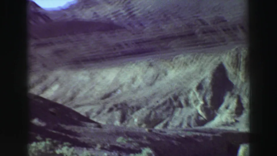 1969 8mm landscape frame with ridged rocky hills, a pale path, and rough foreground terrain, with no people or buildings visible.