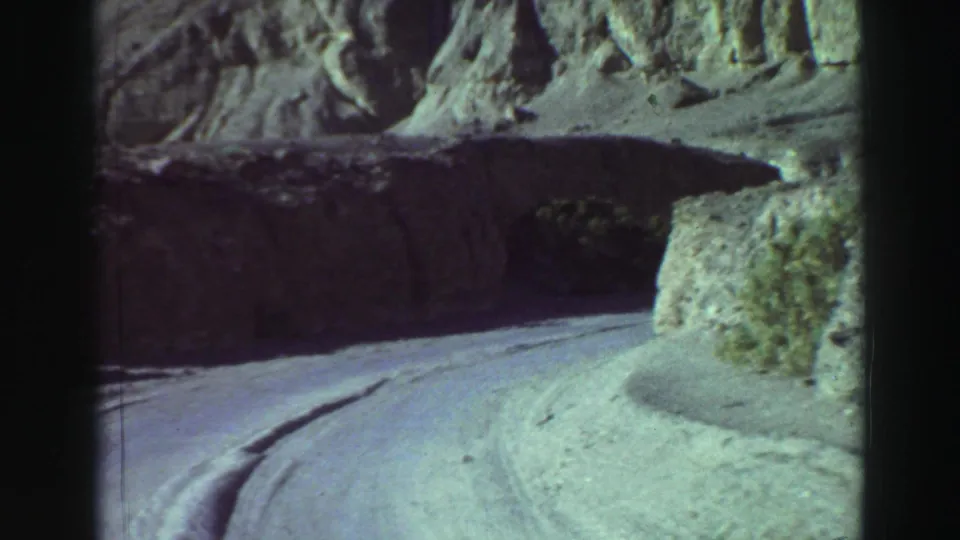 1969 8mm view near Lake Tahoe of a narrow paved road bending between sunlit rock walls and deep shadow.