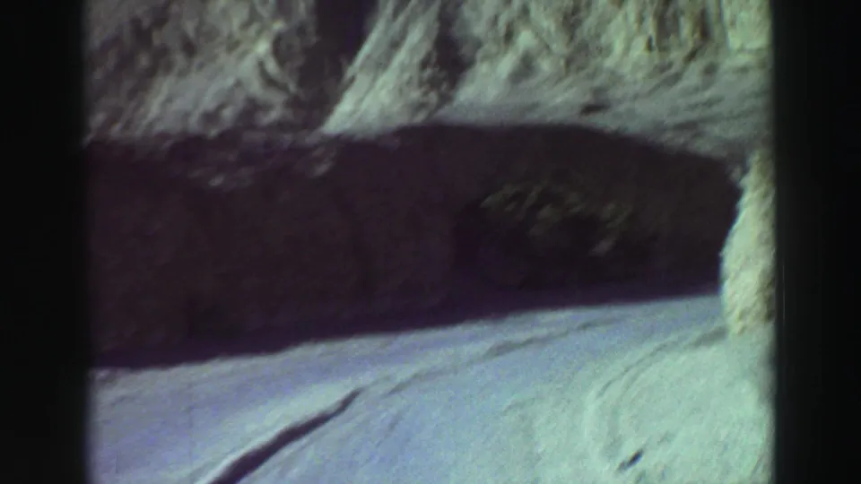 1969 8mm driving view near Lake Tahoe, with a pale road curving past a shadowed cliff and sunlit rocky hillside.