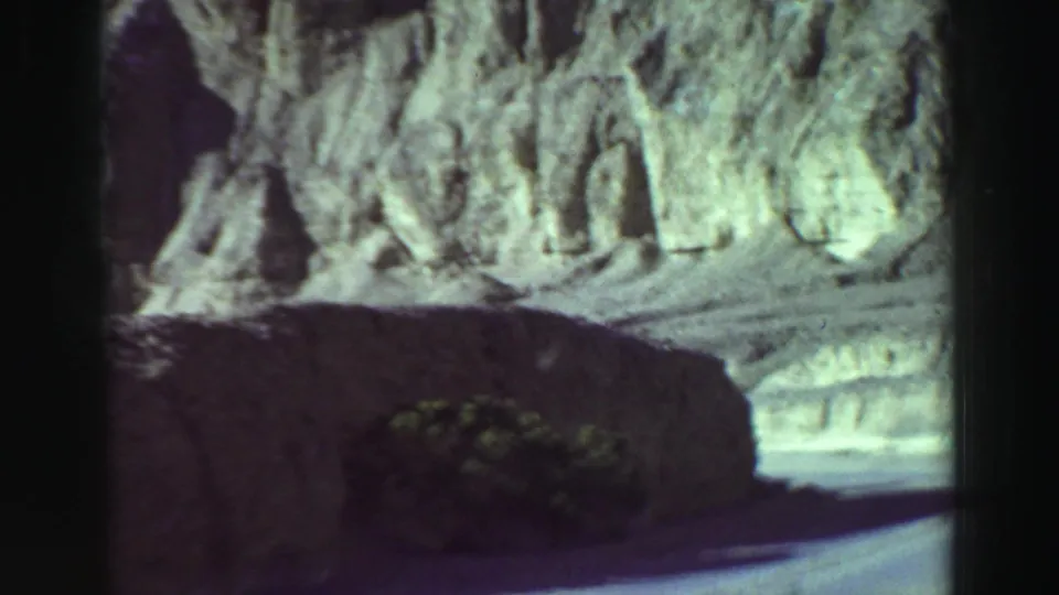 1969 8mm road-trip frame near Lake Tahoe showing eroded rock walls, cliff shadow, scrub, and pavement ahead.