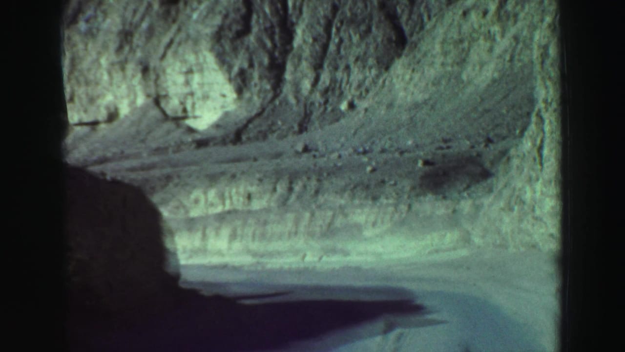 Dirt road curving beside steep rocky slopes seen through a car window.