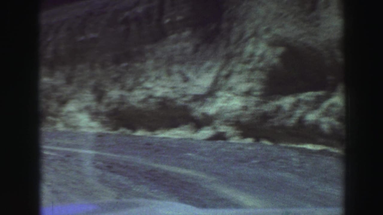 Unpaved mountain road beside a rocky hillside, seen from a moving vehicle.