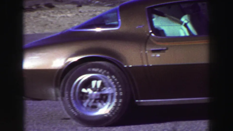 Lake Tahoe 8mm home-movie frame showing the side and rear of a bronze classic coupe with wheel, door, window, and asphalt foreground.