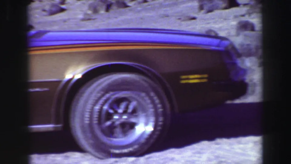 The pan reaches the car's front section, showing a chrome wheel, side stripe and rocky California ground in bright sunlight.