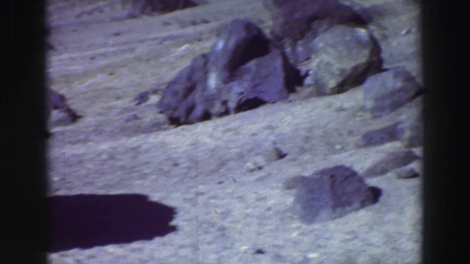 A home-movie frame of sandy terrain and boulders near the parked car, with deep shadow and visible 8mm color grain.