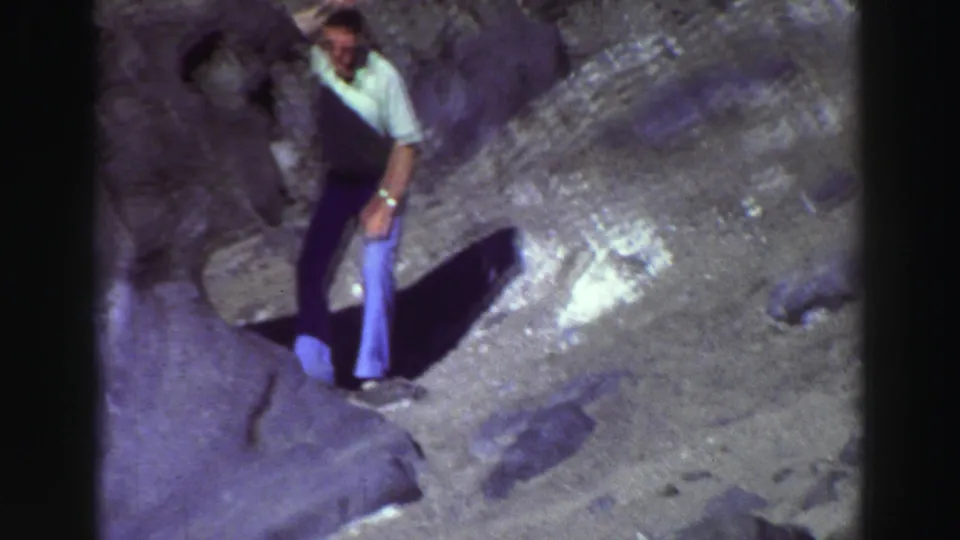 Man in a light polo shirt and flared pants stands on rocky Lake Tahoe terrain in a 1969 8mm home-movie frame.