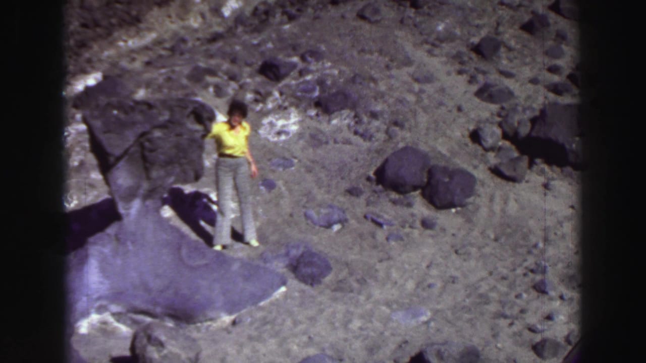 Woman in a yellow shirt stands among dark rocks on dusty ground.