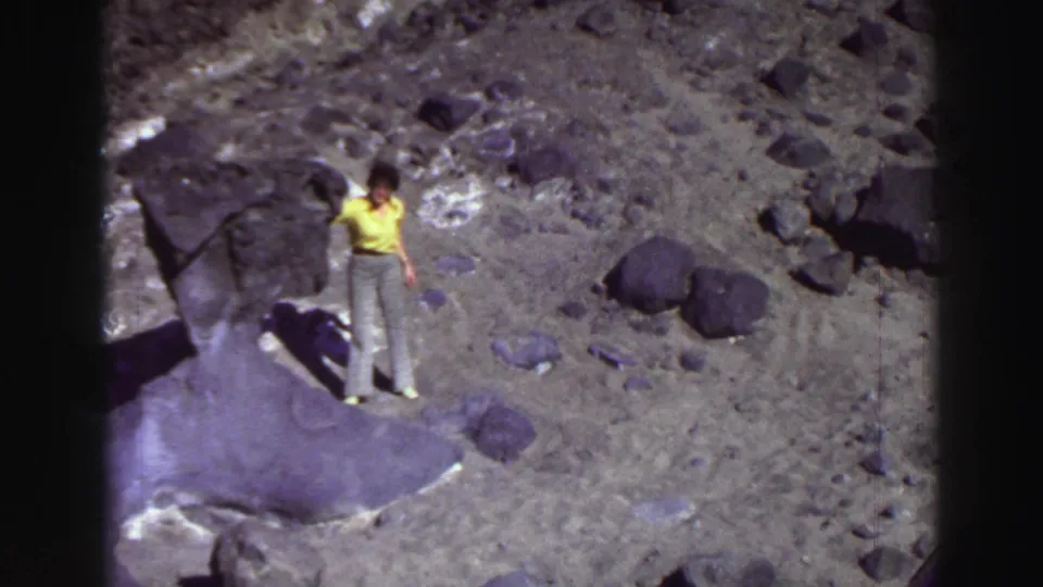 Dark-haired woman in a yellow shirt and light pants stands on rocky Lake Tahoe terrain in 1969 home-movie footage.