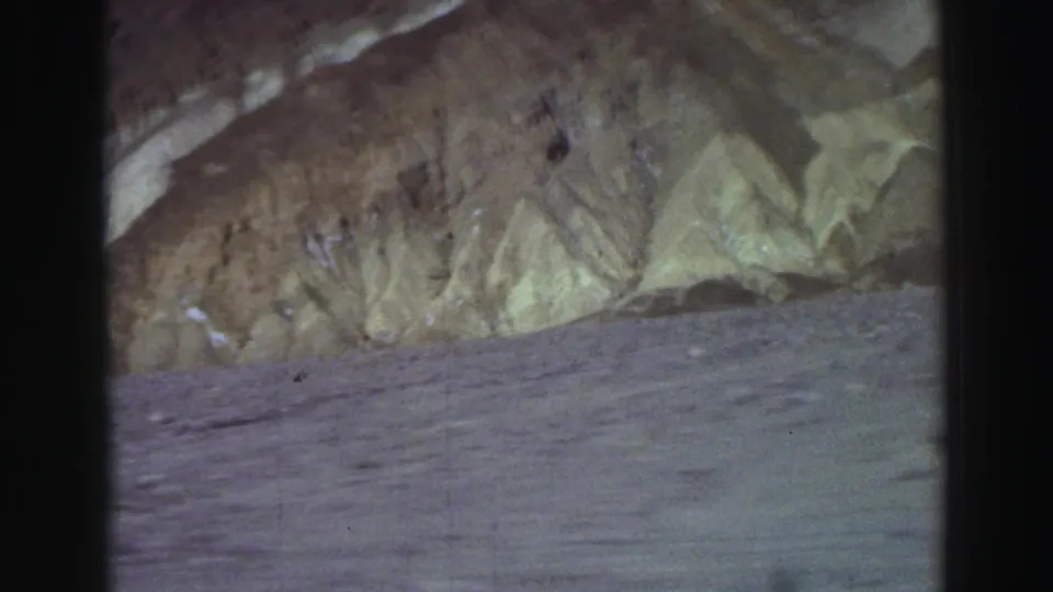 A 1969 8mm frame shows arid ground and rugged mountain layers, partially bordered by dark vertical edges from the camera or vehicle position.