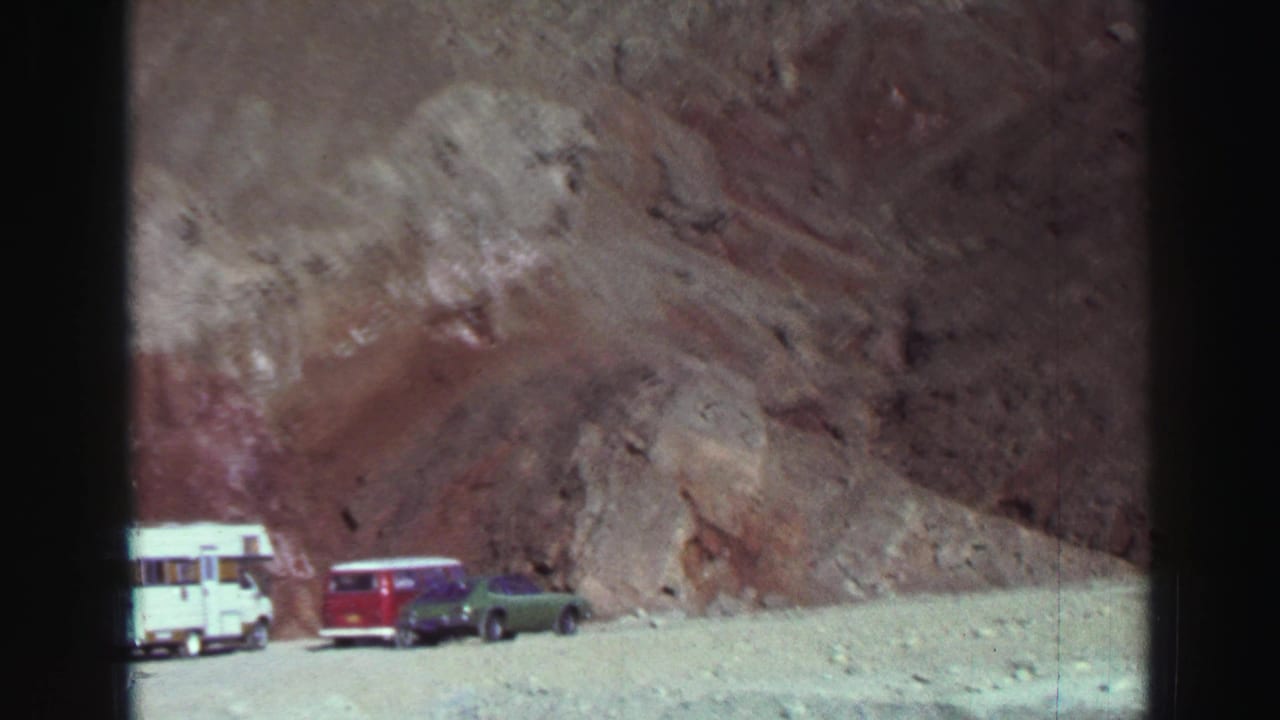 White camper, red wagon, and green car parked below a rocky slope.