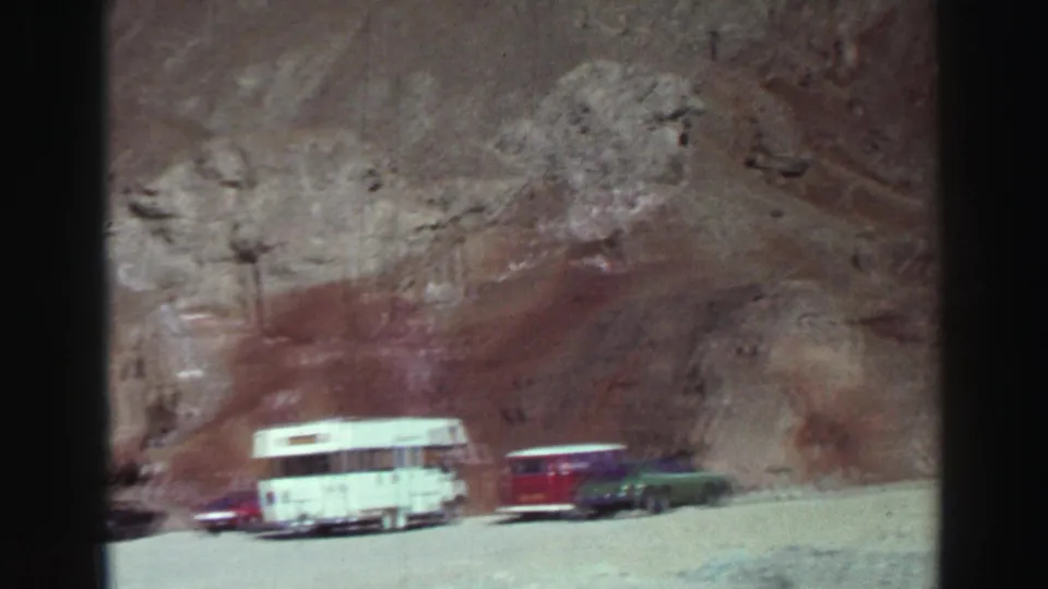 Lake Tahoe 1969 frame showing a white camper, red vehicle, and green car lined up below a sunlit rock formation.