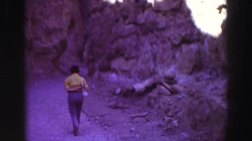 1969 Lake Tahoe home-movie frame with a distant person in a yellow shirt walking through a narrow rocky canyon area.
