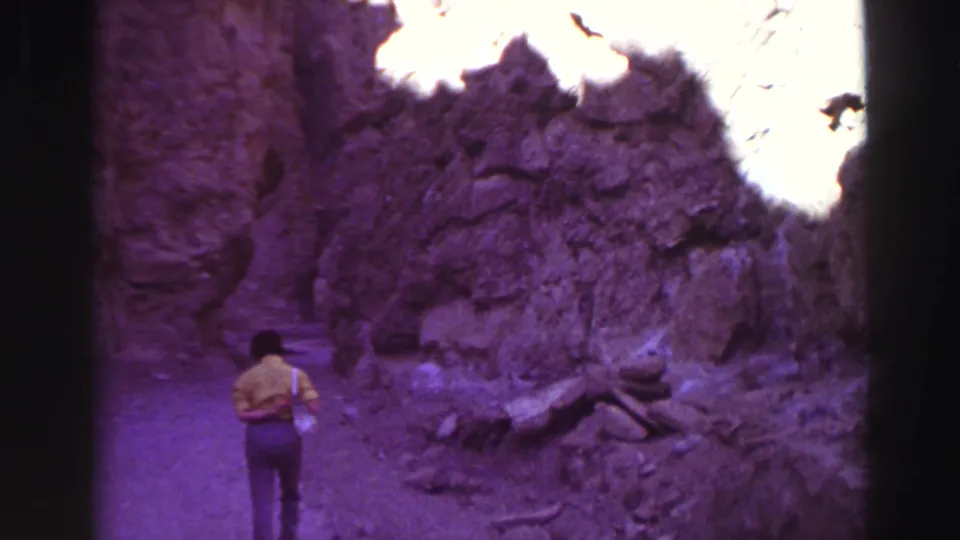 1969 8mm home-movie frame near Lake Tahoe showing a lone walker entering a shadowed rocky canyon with bright sun on the cliffs.