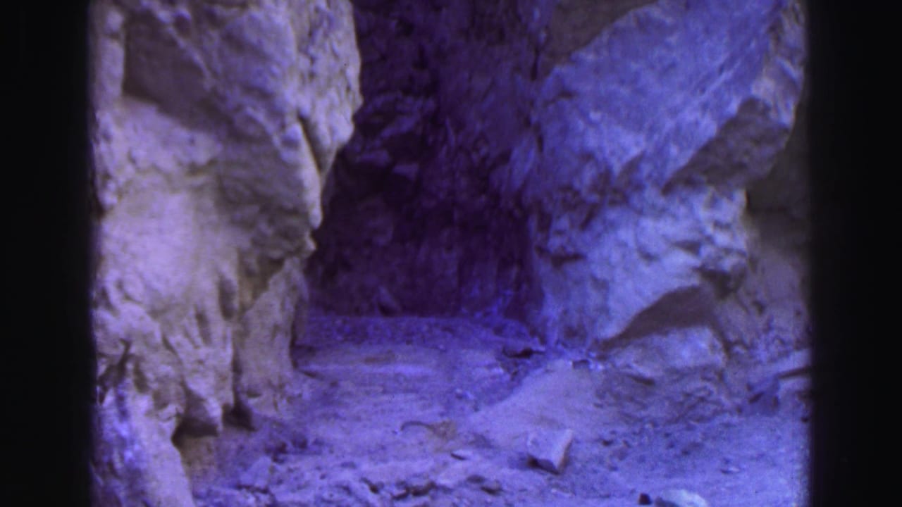 Narrow rocky passage with dirt ground and purple-blue film cast.