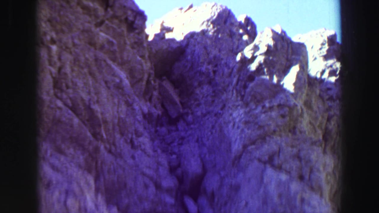 Rocky cliff wall rising toward jagged sunlit peaks under blue sky.
