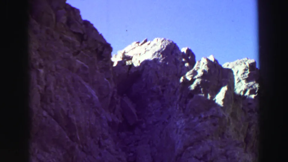 1969 Lake Tahoe home-movie frame with rough cliff textures, bright ridge tops, and a narrow band of blue sky.