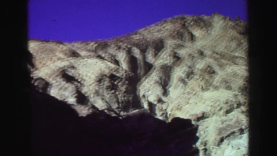 1969 8mm Lake Tahoe landscape frame with a rocky ridge, shadowed foreground canyon wall, and saturated blue sky.