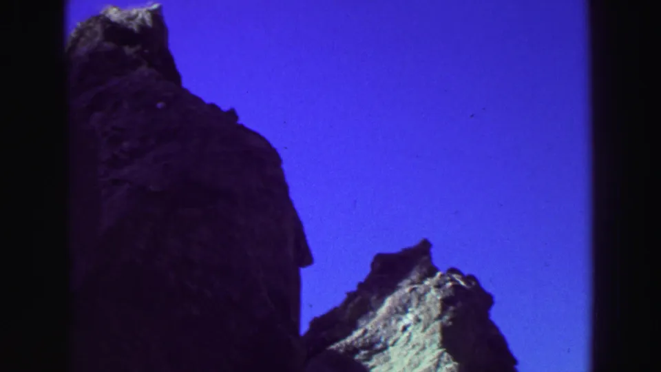 1969 Lake Tahoe 8mm frame showing a tall dark rock formation, smaller sunlit stone peak, and open blue sky.
