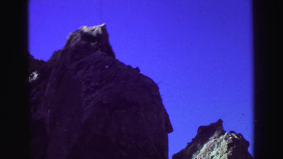 1969 Lake Tahoe home-movie frame with a large shadowed rock face, a small sunlit outcrop, and saturated blue sky.