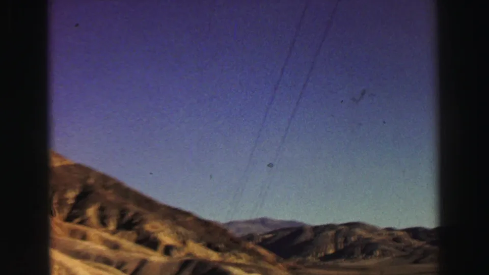 1969 Lake Tahoe-area home-movie view of a dry hillside, distant ridges, blue sky, and faint utility lines from a moving car.