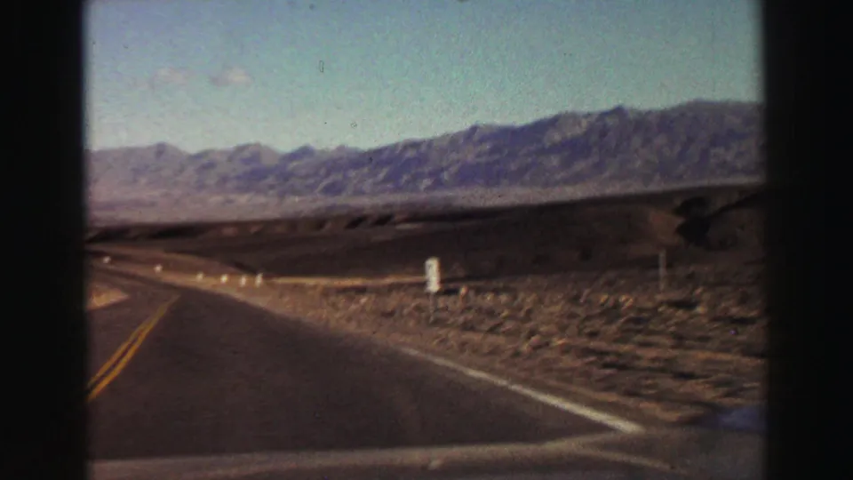 1969 Lake Tahoe-area driving frame showing a two-lane road, roadside posts, dry valley floor, and distant mountain range.
