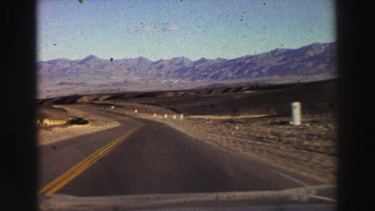 Two-lane road curving through dry terrain toward distant mountains.