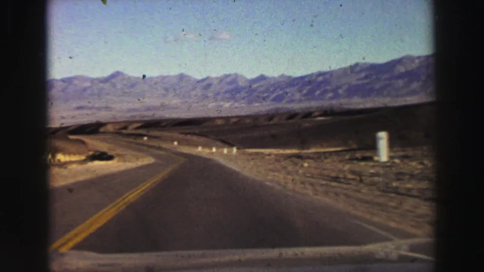 1969 8mm travel view from a moving car on a paved road through arid terrain near Lake Tahoe, with mountains in the distance.
