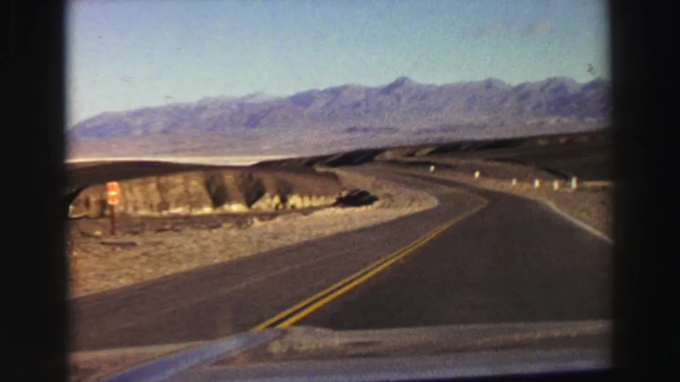 A 1969 home-movie frame shows the road bending right past eroded desert banks, with mountains beyond under a clear sky.