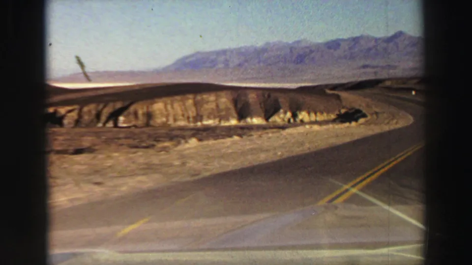 Passenger-view 8mm footage shows a mountain road curve, dry roadside cut, and a faint car windshield edge in the foreground.