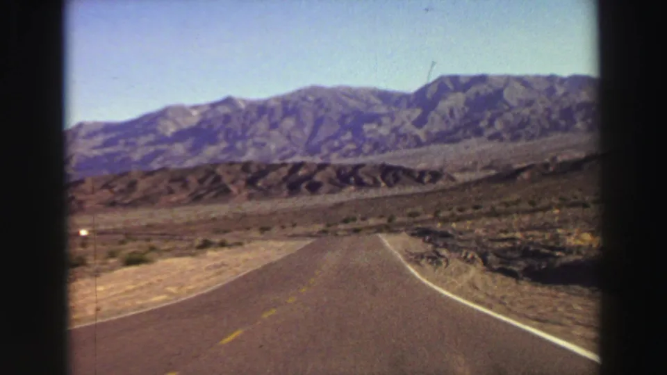 The road straightens toward rugged mountains in this 1969 8mm moving-car view near Lake Tahoe, with open dry land on both sides.
