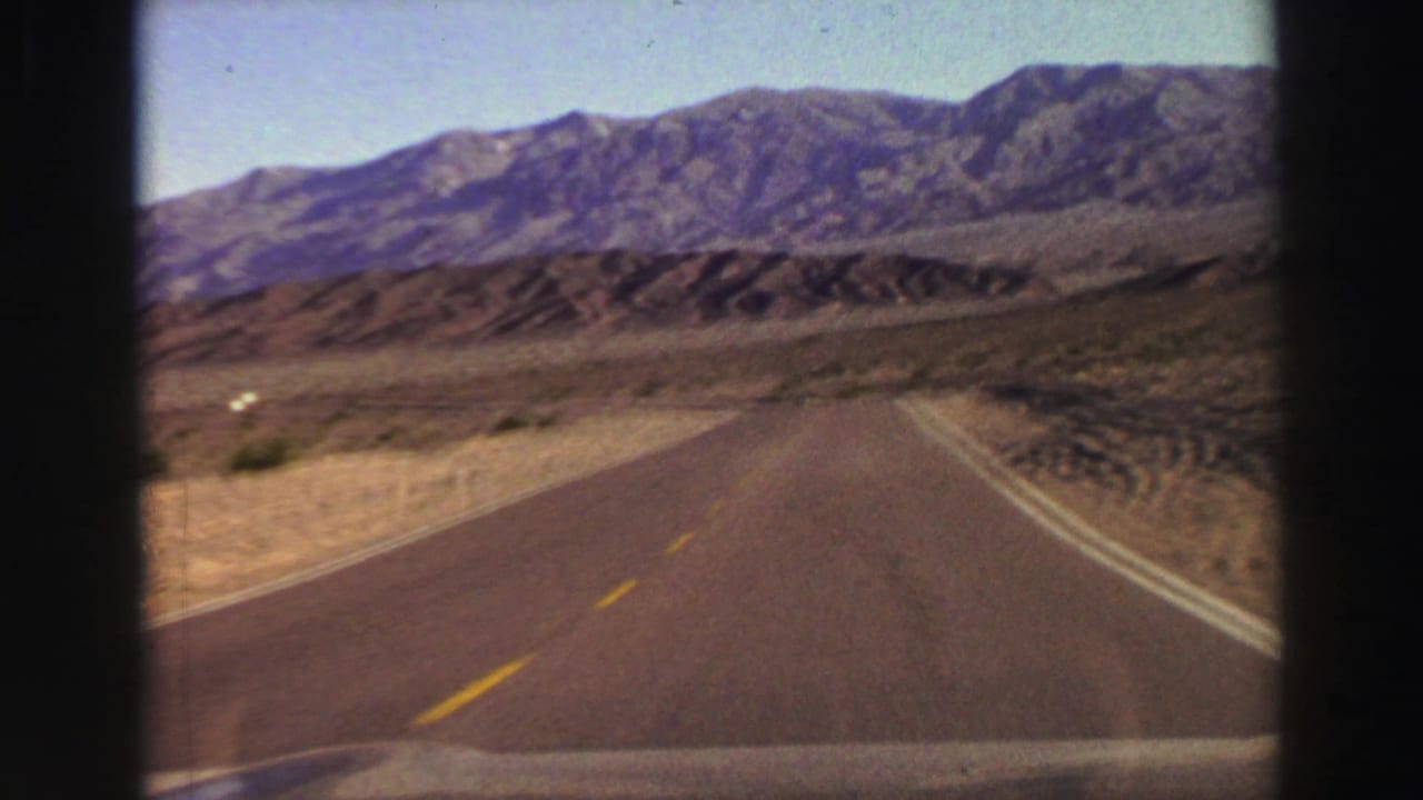 Two-lane road leading toward distant mountains in dry terrain, framed by dark car-window edges.