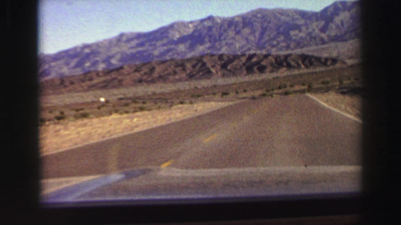 Curving road seen over a car hood with dry roadside land and mountains ahead.