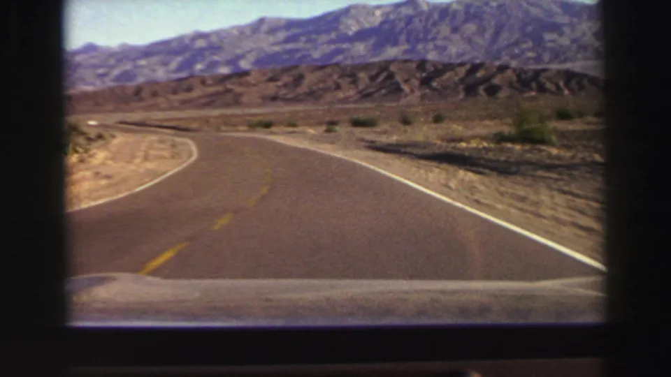 1969 home-movie frame showing a paved road curving through dry California terrain beneath a mountain backdrop.