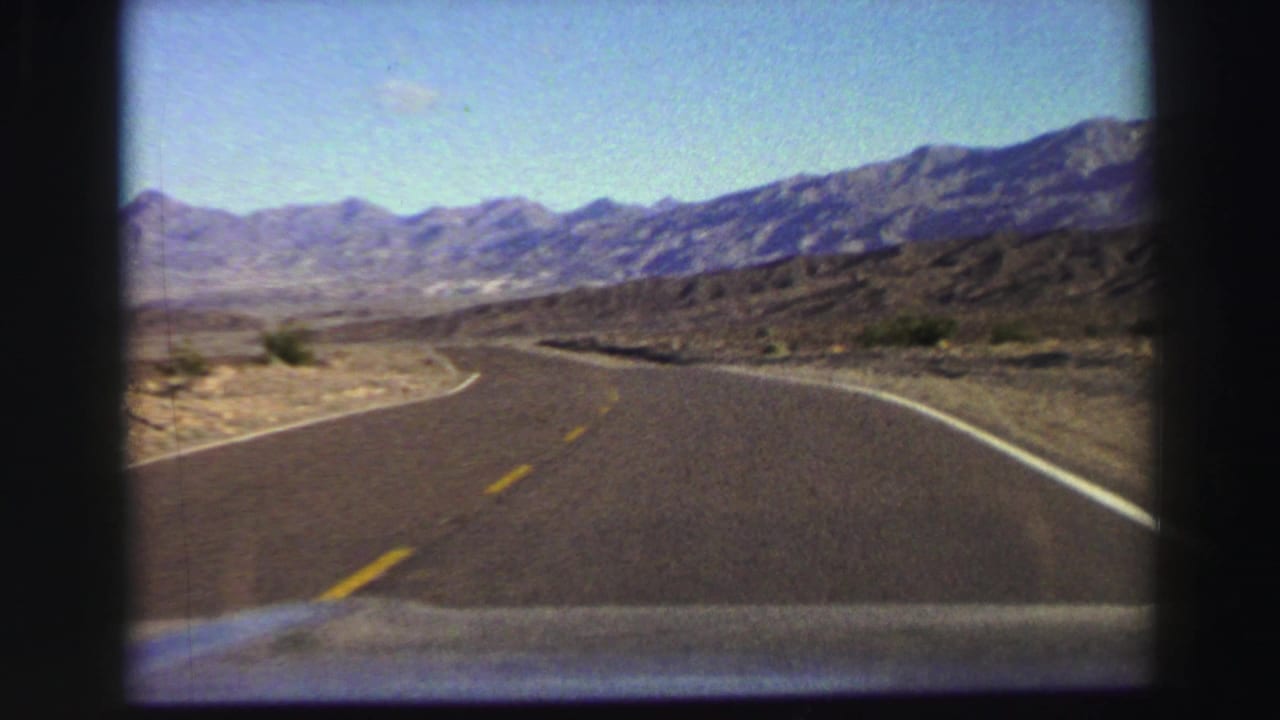 Forward car view of a winding road through dry terrain and mountains.