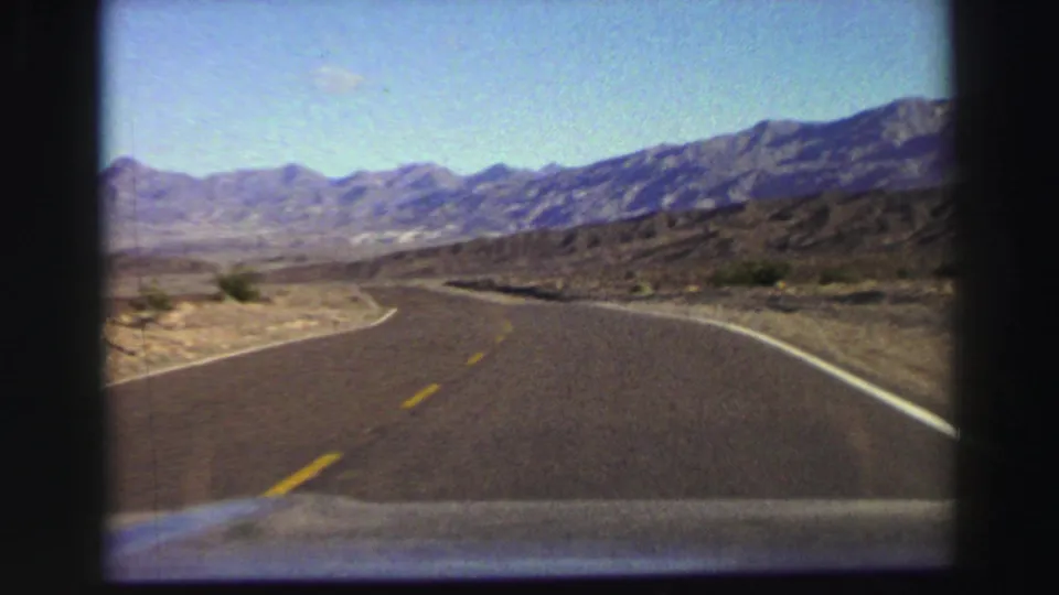 Vintage 8mm road-trip frame showing a paved desert highway bending through scrub toward mountain ridges.