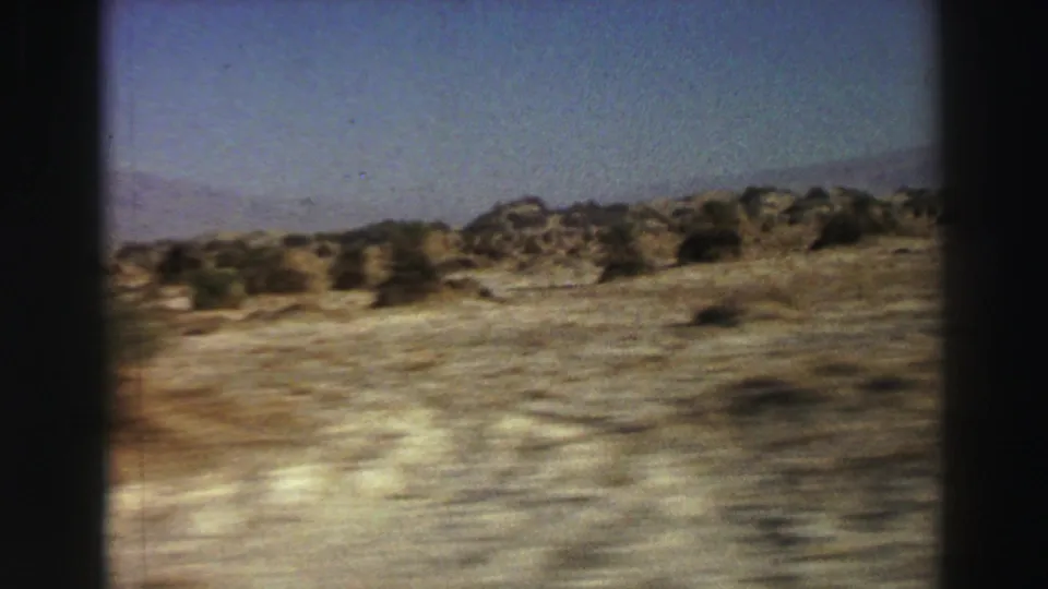 1969 8mm frame of roadside desert passing quickly, with shrubs, rocky soil, and low hills under a clear sky.
