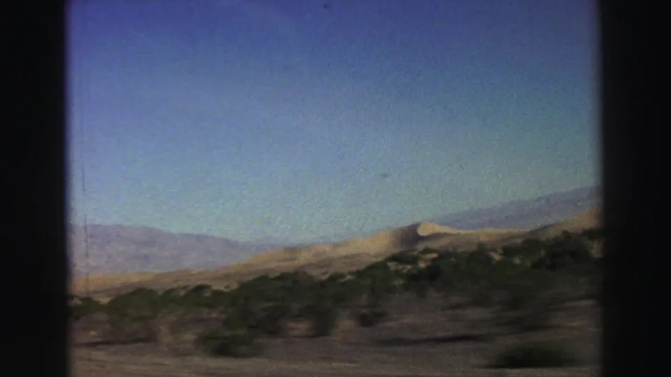 1969 8mm car-window view with a distant mountain backdrop, dry roadside vegetation, open sky, and visible film-frame borders.