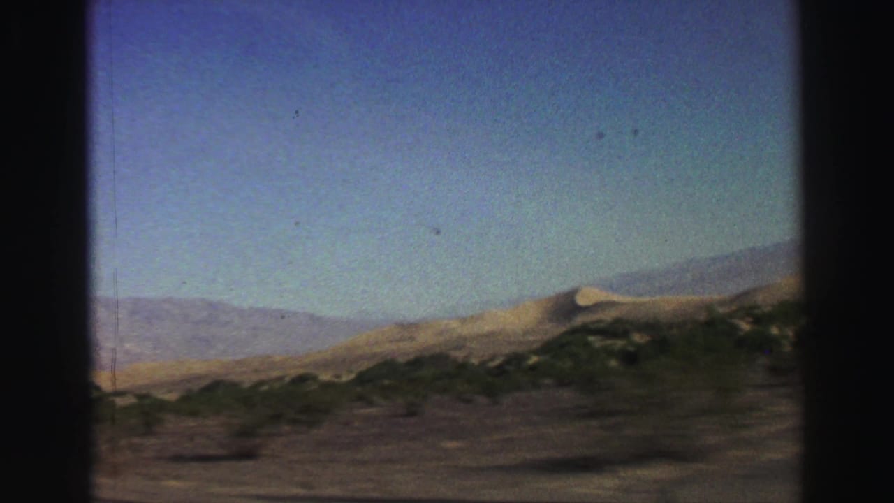Dry hills, shrubs, and distant mountains seen through a dark vehicle-window frame.
