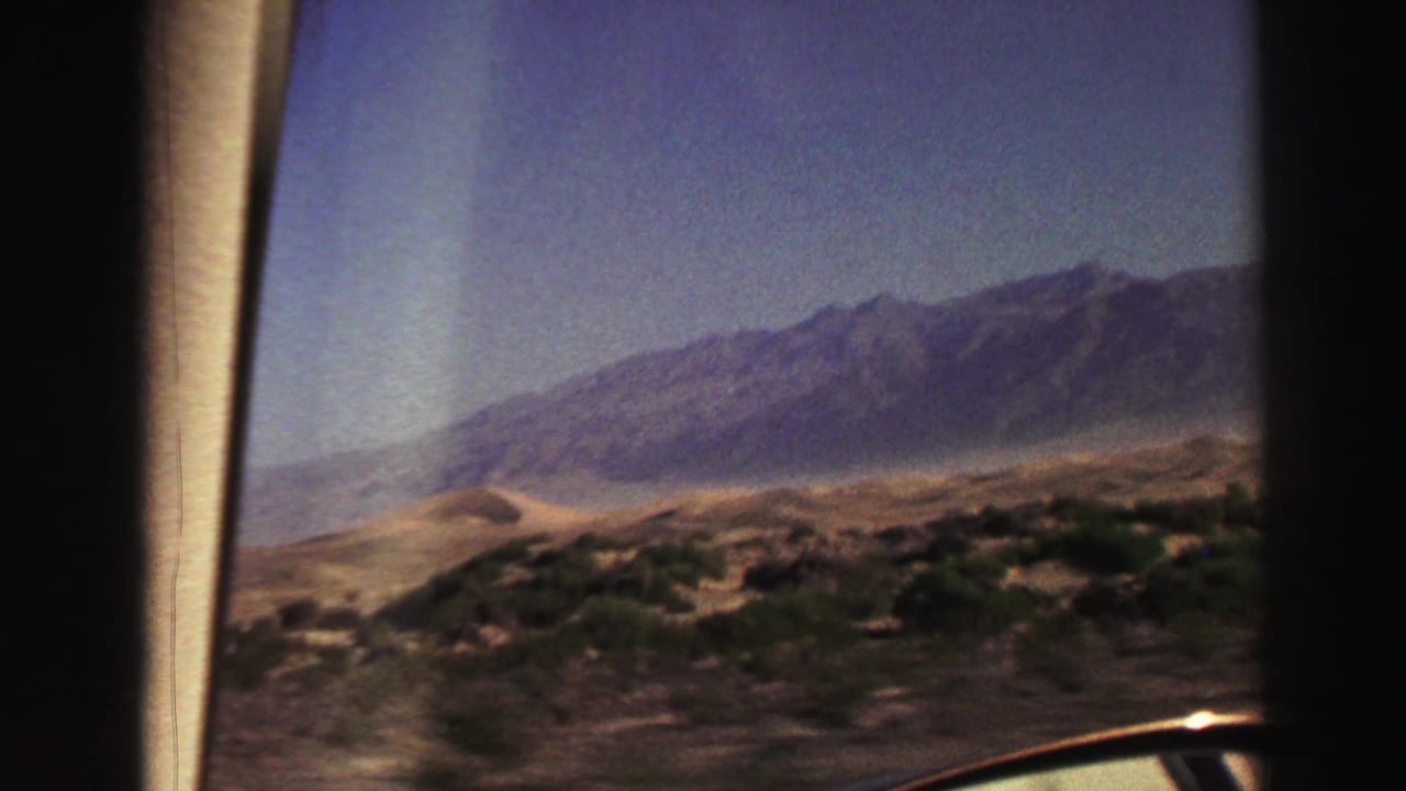View through a car window toward desert scrub, sandy flats, and a distant mountain range.