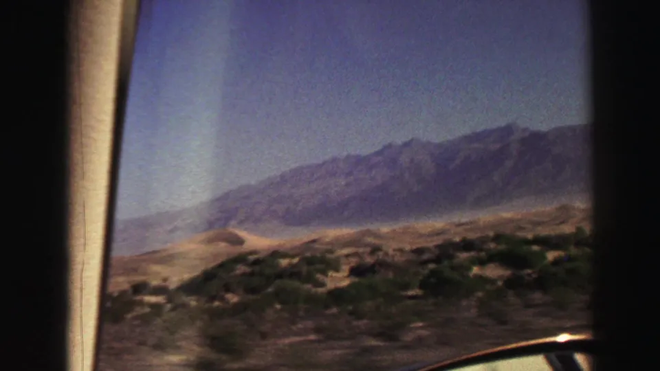 1969 8mm travel view through a vehicle window, showing arid foreground scrub and distant mountains under a blue sky.