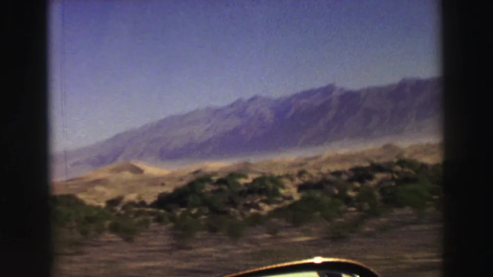 1969 8mm roadside view with a dark vehicle-window divider, low desert vegetation, pale sand, and mountains in the distance.