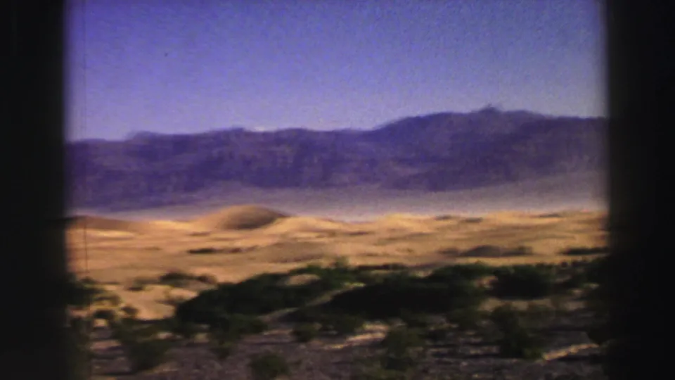 1969 8mm moving-car view of scrubby arid ground, pale sand, and a long mountain ridge beneath a clear sky.